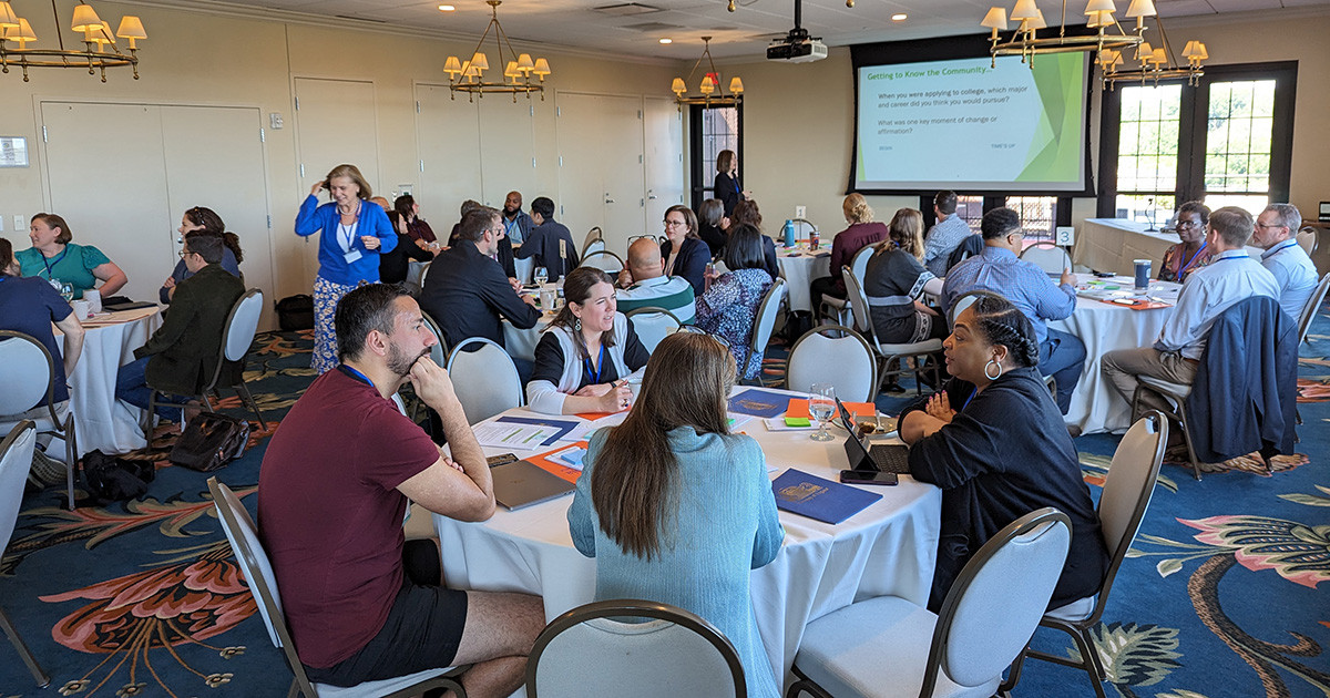 A large group of workshop participants sitting around round tables while talking to each other.