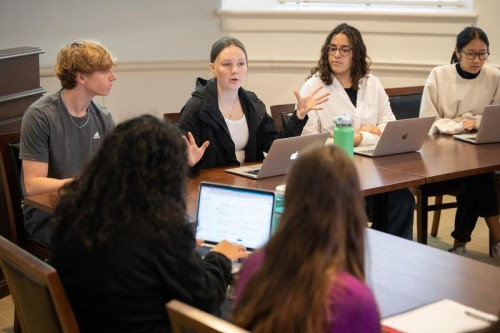 A group of seated students with one classmate gesturing with both hands.