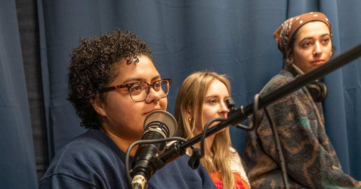 Three female students sit in front of a microphone in a sound studio.