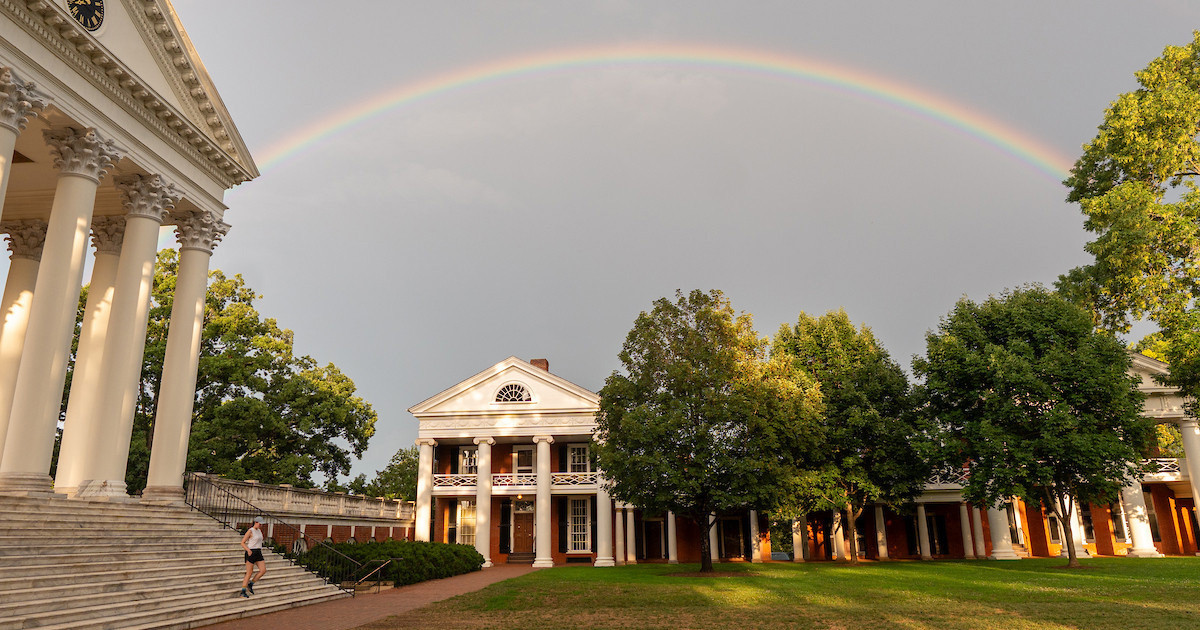 Rainbow arches over a sunlit lawn and neoclassical campus buildings with white columns, framed by trees.