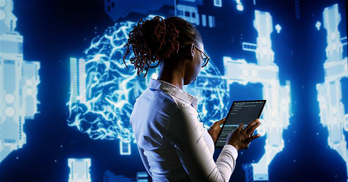 A female facing a wall-sized screen while holding an electronic tablet.
