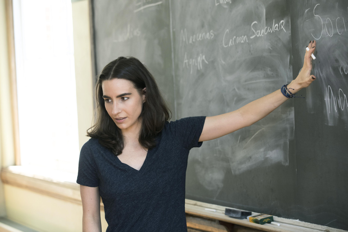 Female Professor holding a piece of chalk and leaning against a blackboard