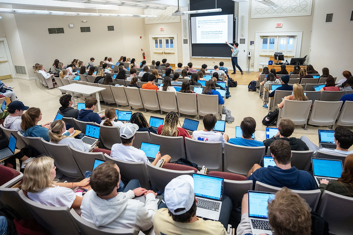 Large classroom of students seated with laptops as professor teaches in the front of the room.