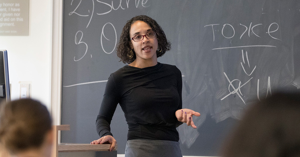 A teacher gesturing to students as she stands in front of a chalkboard.
