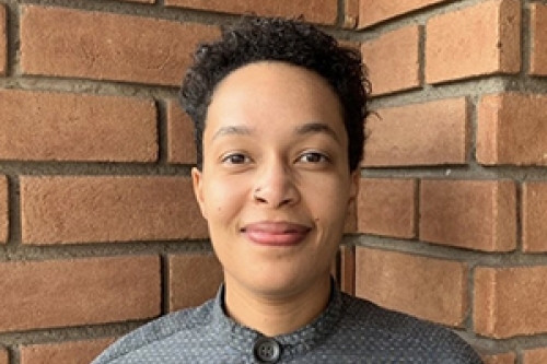 Headshot of Kat Cosby wearing gray shirt standing in front of a brick wall