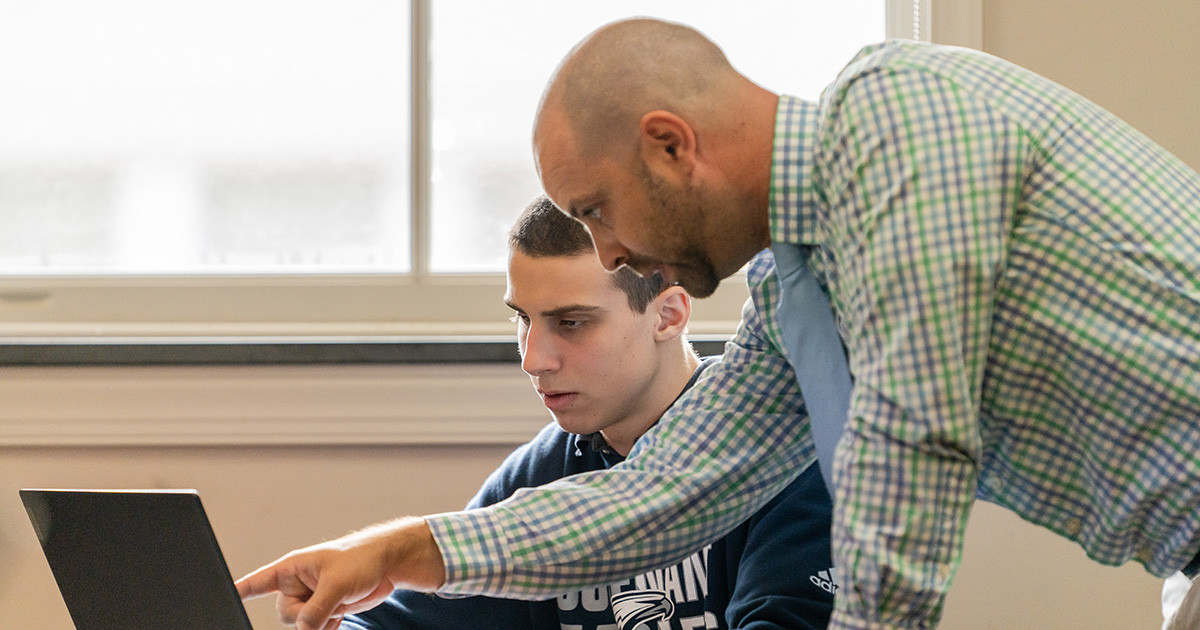 A teacher directing a student who is sitting at a table, working on his laptop.
