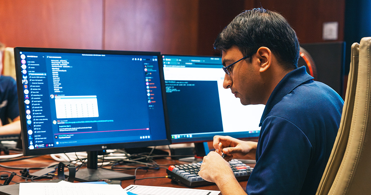 A person sits in front of a computer monitor while looking at a document laying on the desk.