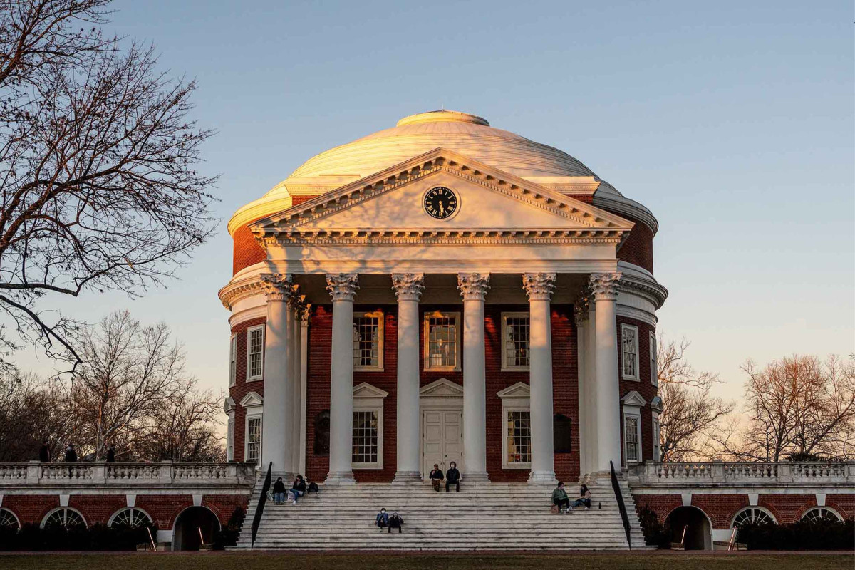 The UVA Rotunda building pictured in the Fall at sunset. There are a few students scattered around, sitting on the steps.