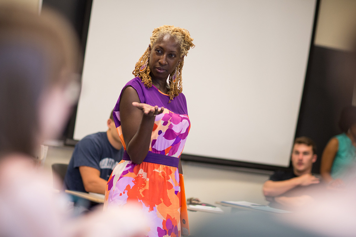 A female professor is standing and gesturing in the middle of a group of seated students.