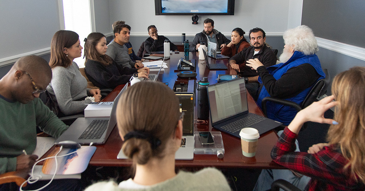 A group of adults sitting at a conference table with laptops, while having a discussion.