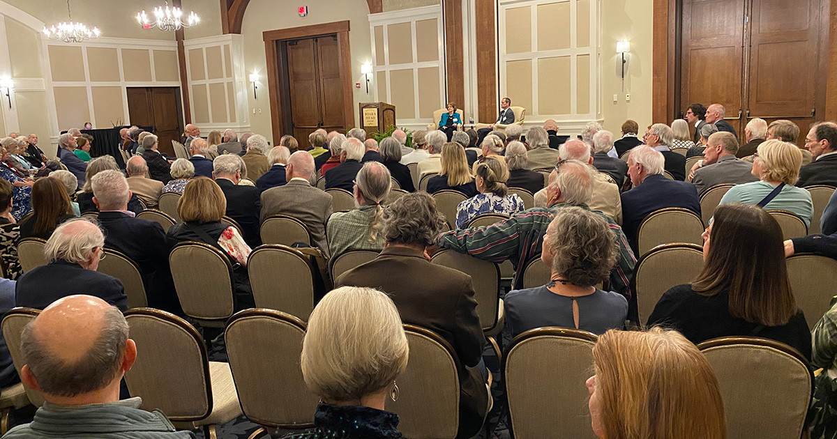 A large audience sits in a large banquet room while listening to a lecture.