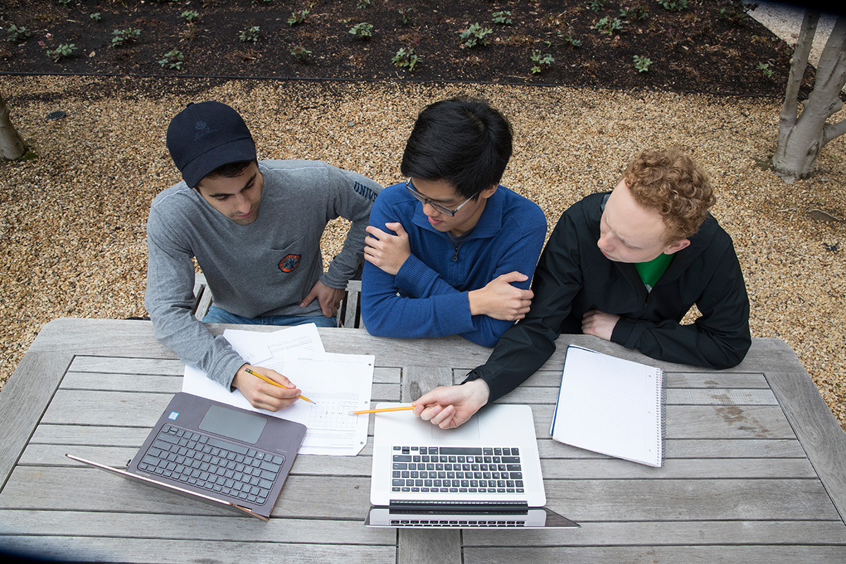 Three students sitting at a table outside while looking at each others notes.