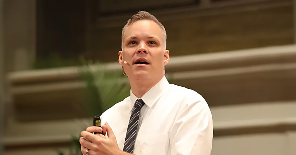 Ryan White, wearing a white shirt and tie with a headset mic, speaks while holding a presentation remote in an indoor lecture space.