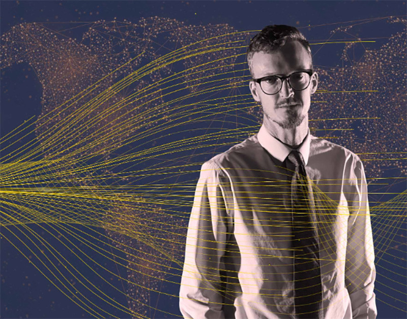 Dark image of a man dressed in a shirt and tie stands in front of a map of the world with lines representing data flows over images.