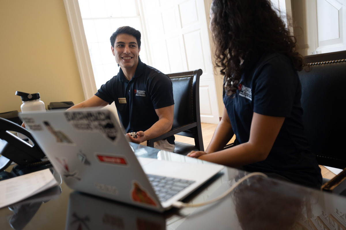 A man and woman sitting at a table, talking while they use their laptops.