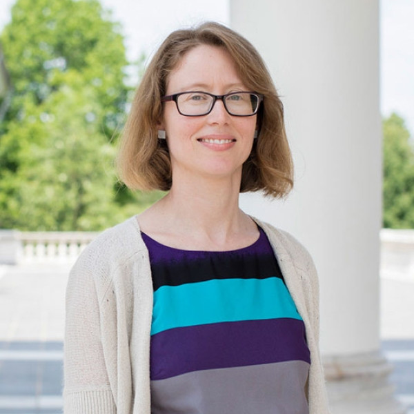 Headshot of Abby Self with short, light-colored hair wearing a striped top with a light cardigan, standing outdoors near white columns.